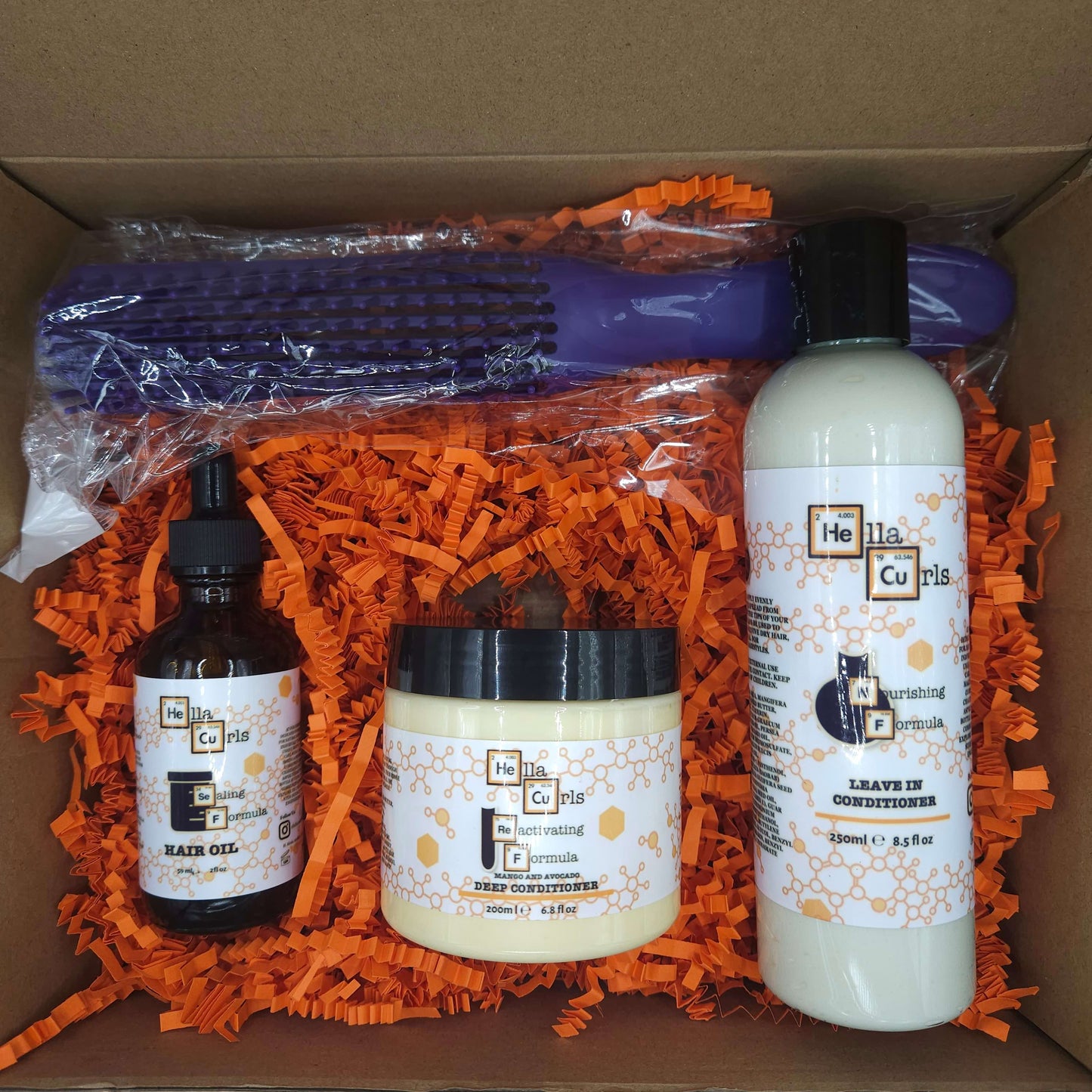 Set of hair care products including a bottle, jar, and brush in a box with orange shredded paper.